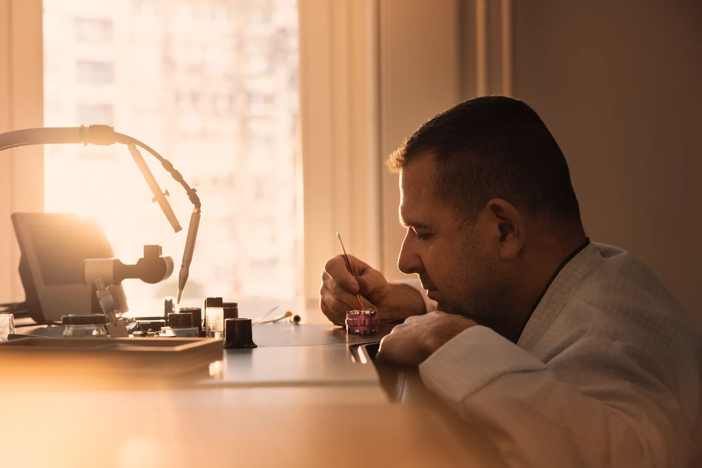Man carefully working on a small mechanical watch component at a workbench with soft sunlight in the background.