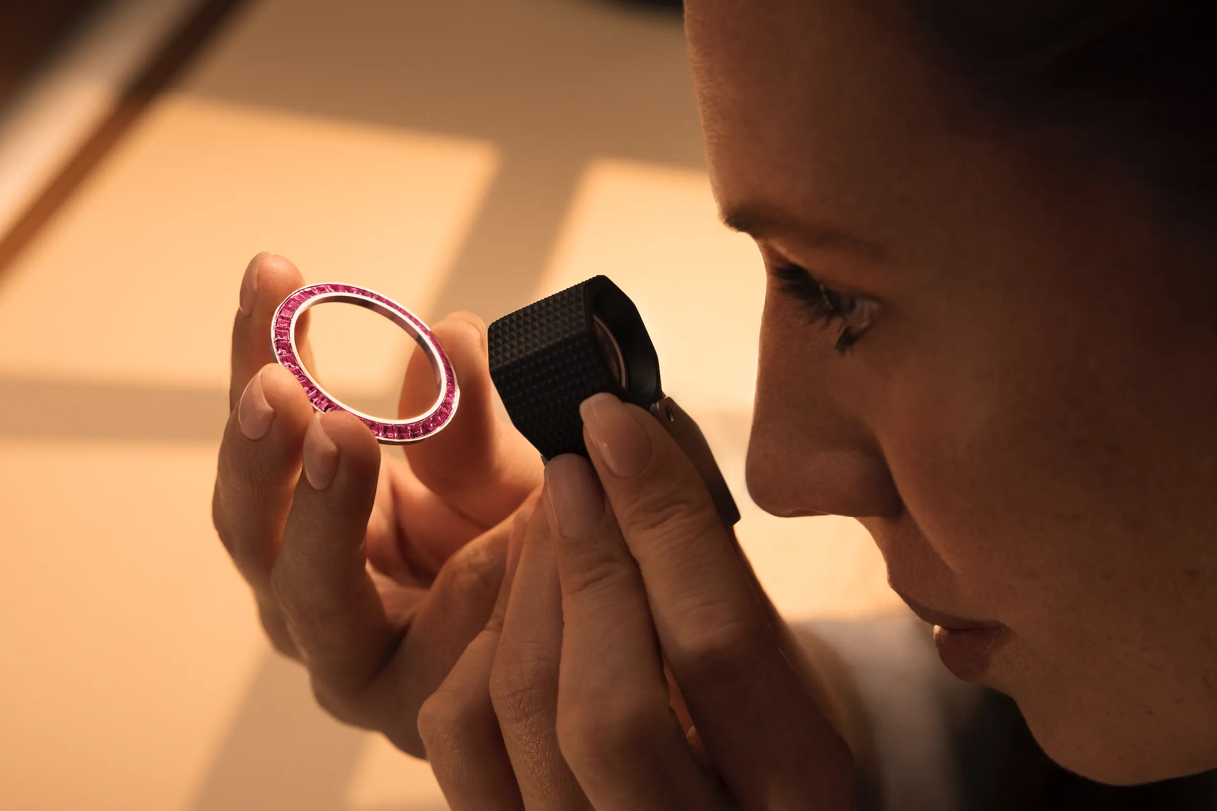 Close-up of a woman inspecting a pink gemstone bezel with a jeweler's loupe.