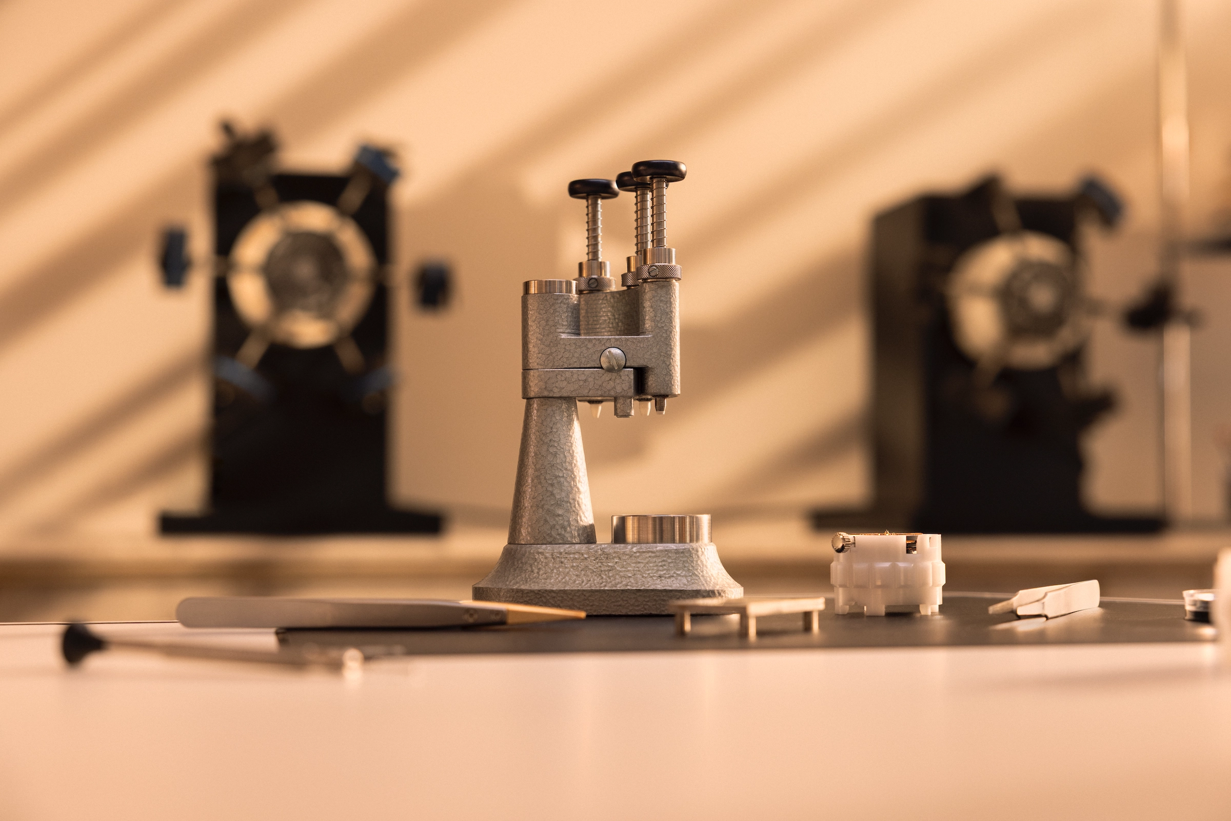 Close-up of a precision watchmaking tool with miniature parts on a workbench and blurred watchmaker devices in the background.