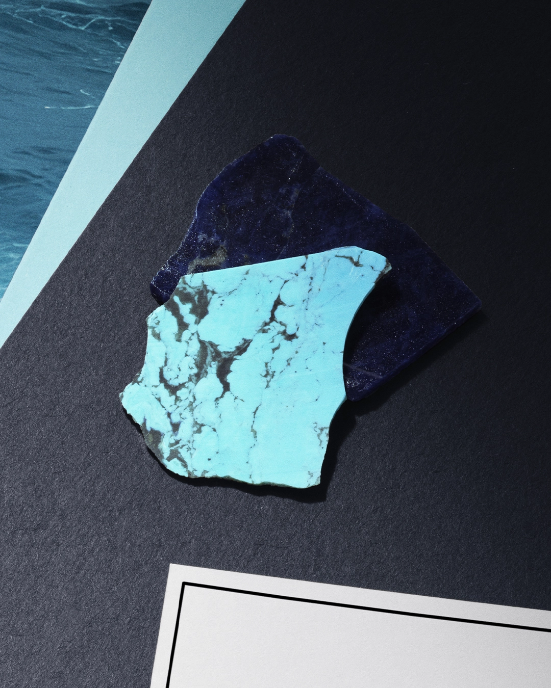 Two irregularly shaped stone pieces, one turquoise with black veins and the other dark blue, placed on a dark textured surface.