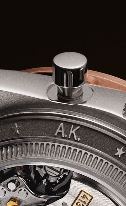 Close-up of a watch crown and part of the watch mechanism showing engraved initials 'A.K.' on the metal surface.