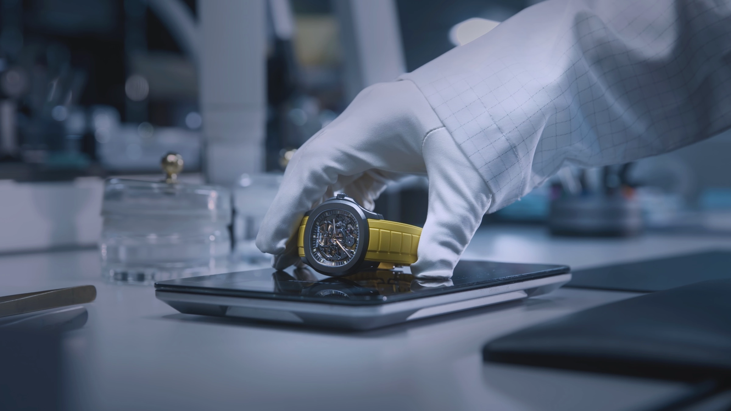 Gloved hand placing a watch with a yellow strap on a digital scale in a lab setting.