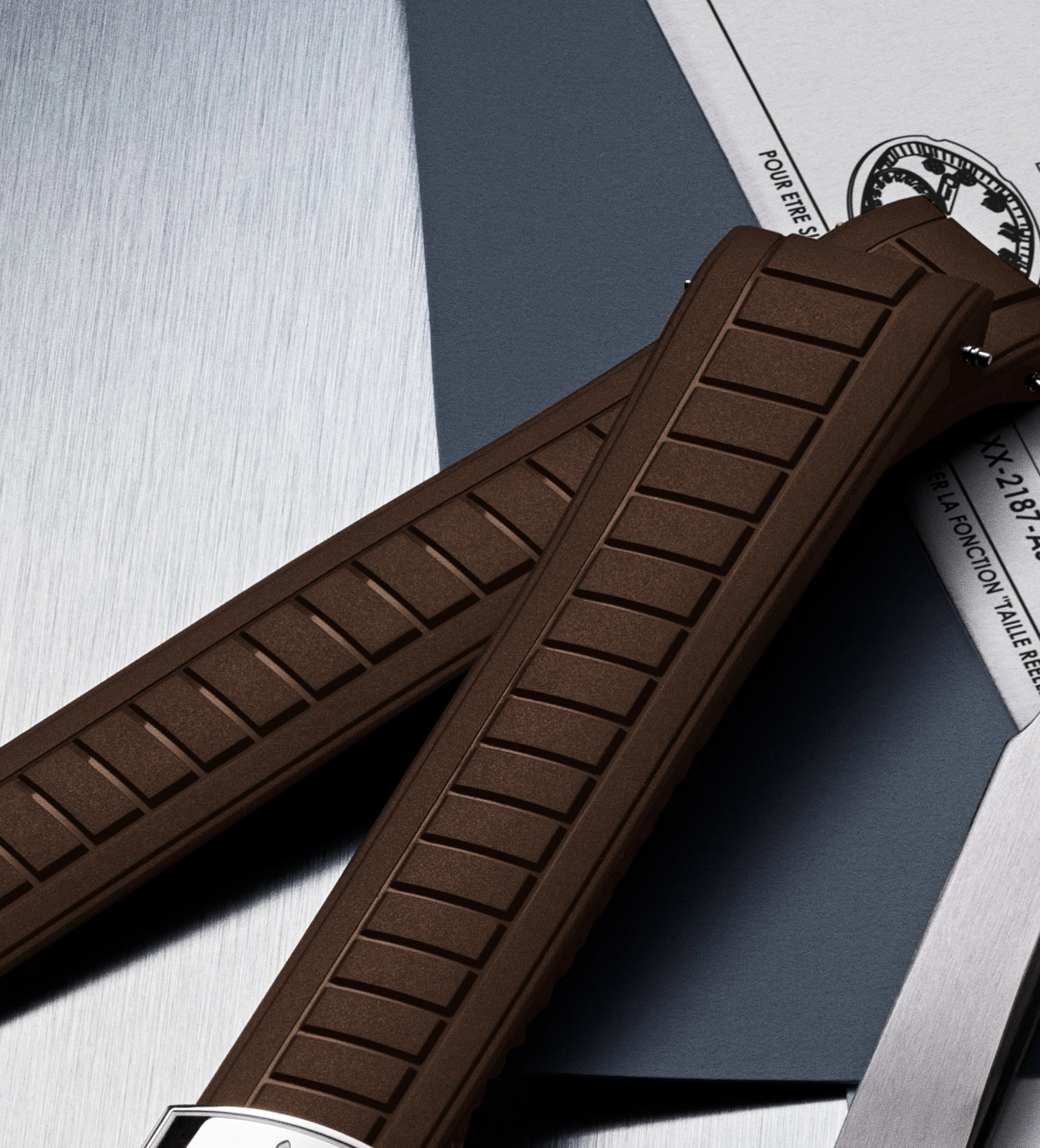 Close-up of a brown textured watch strap with metal clasp resting on documents and a brushed metal surface.
