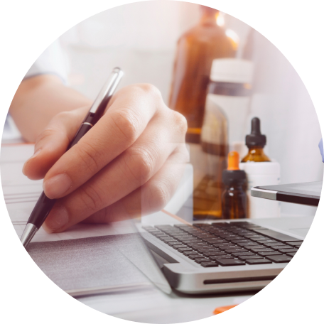 Hand holding a pen poised over paper with a laptop keyboard and medicine bottles in the background.