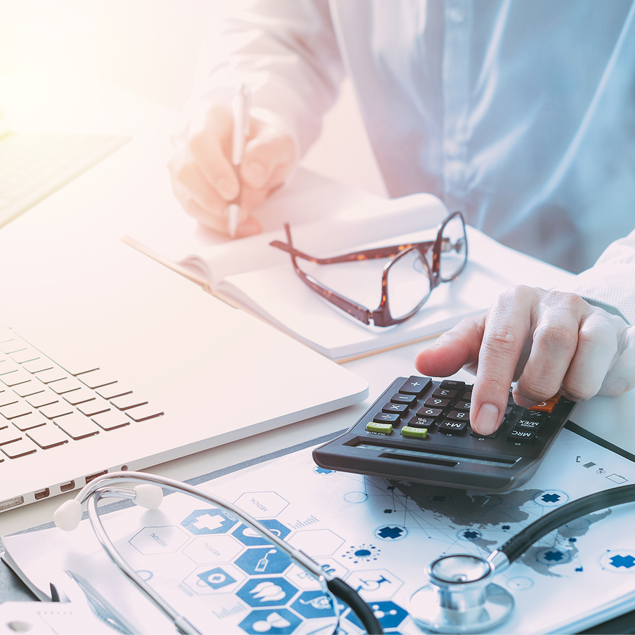 Person using a calculator on a medical-themed desk with a laptop, stethoscope, glasses, and notebook.