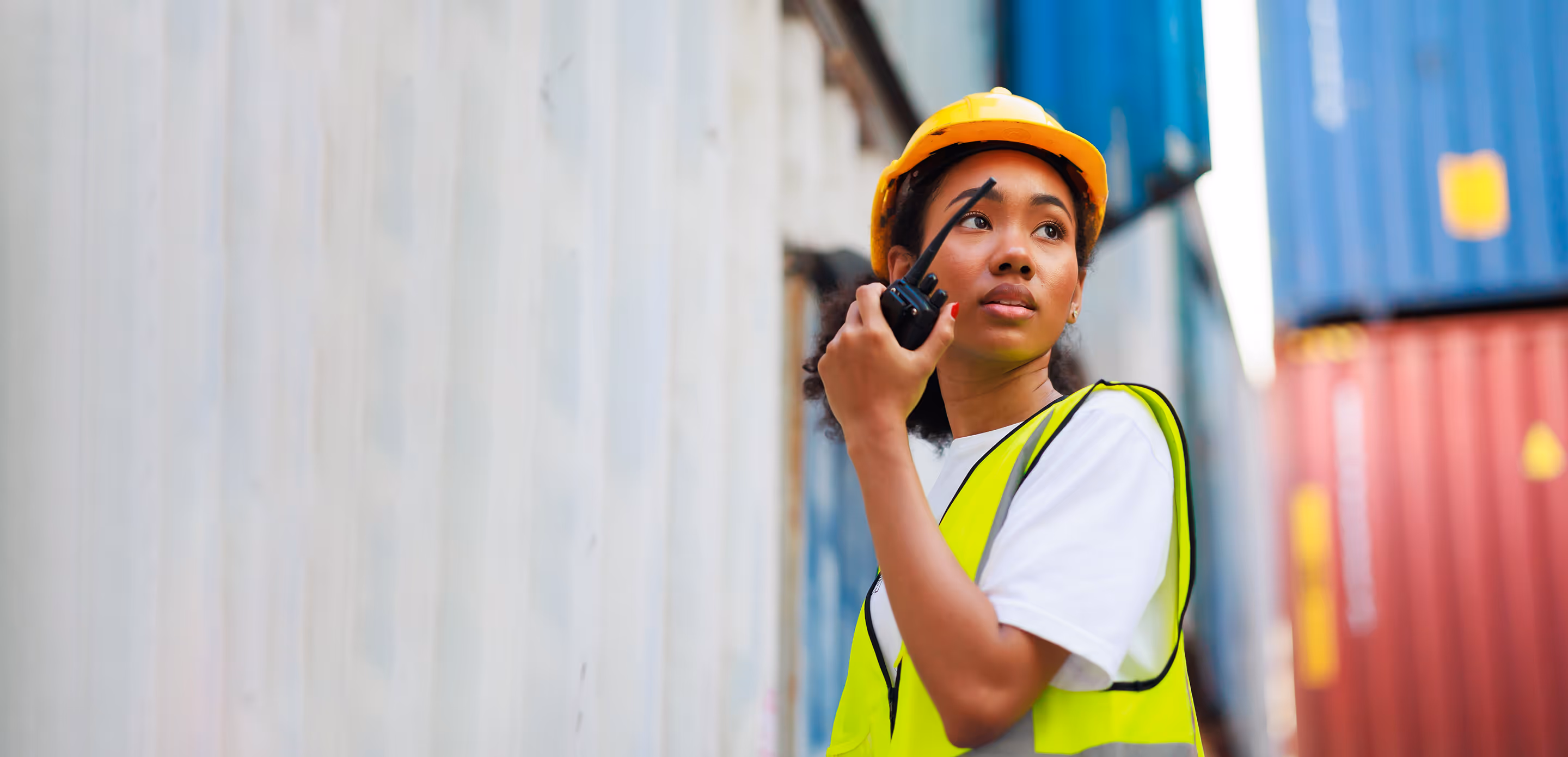 Woman wearing a yellow hard hat and safety vest communicating on a walkie-talkie near shipping containers