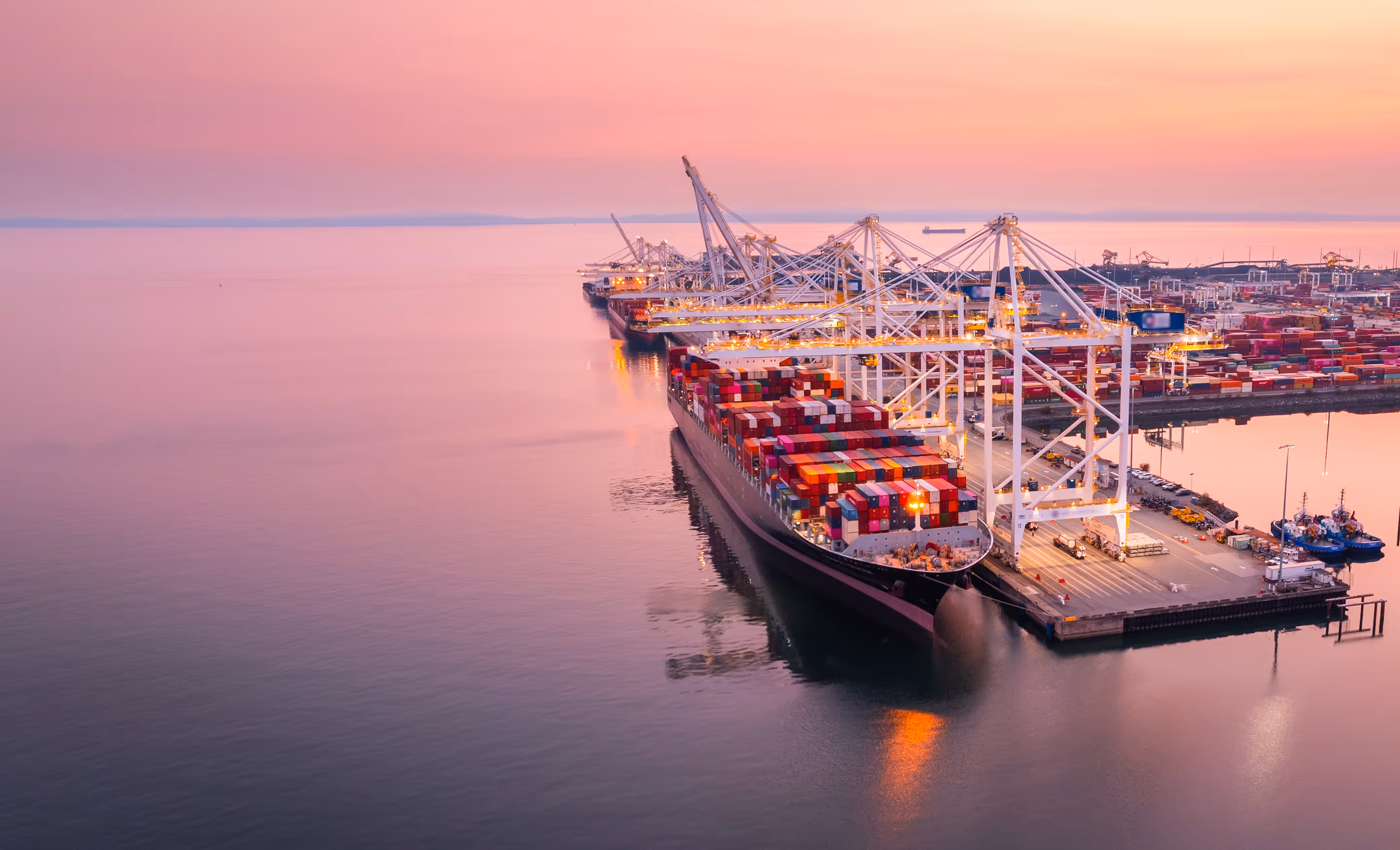 Large container ship docked at a busy maritime port at sunset, with cranes loading cargo and calm water reflecting the lights.
