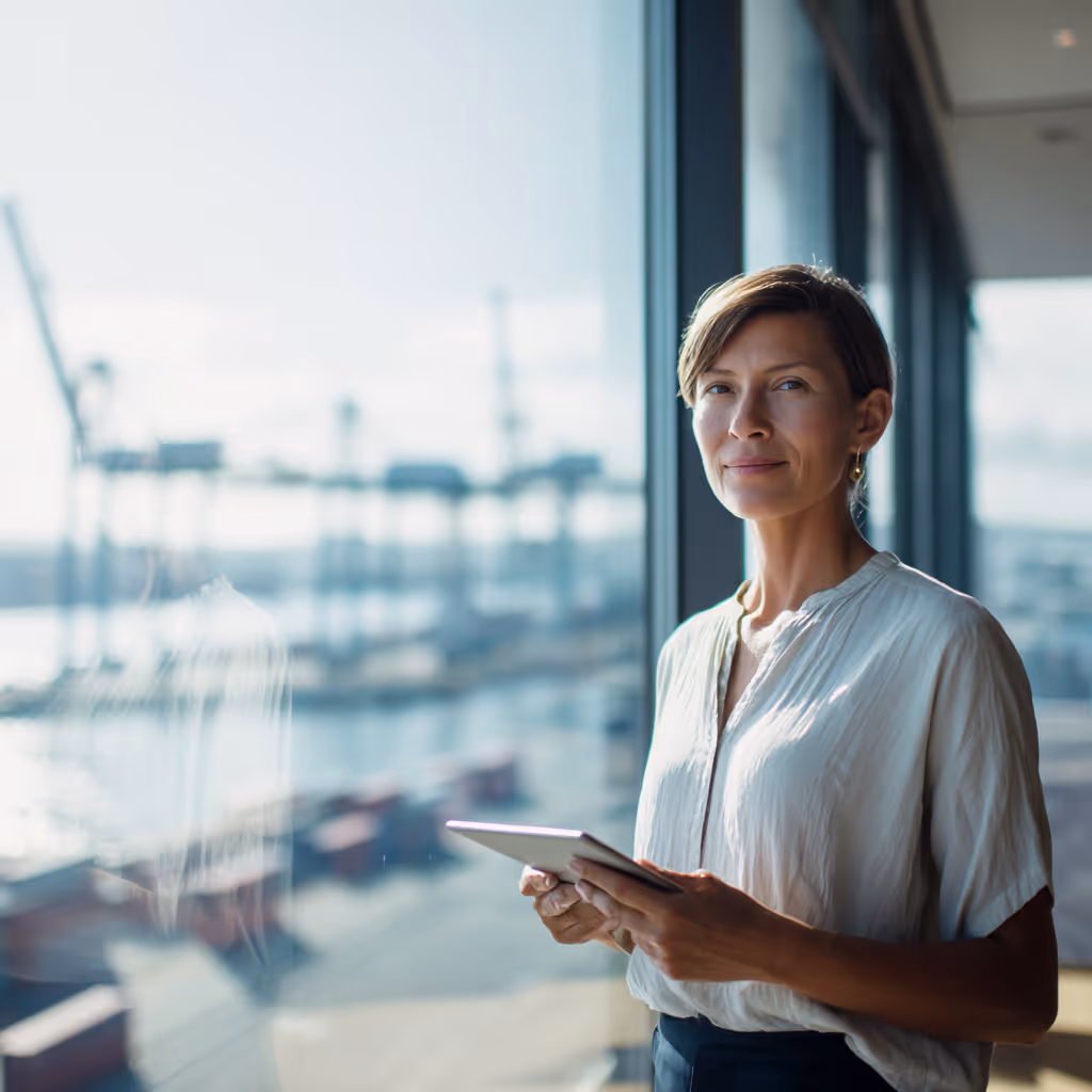 Female operations manager holding a tablet while standing by a window overlooking a shipping port and container cranes