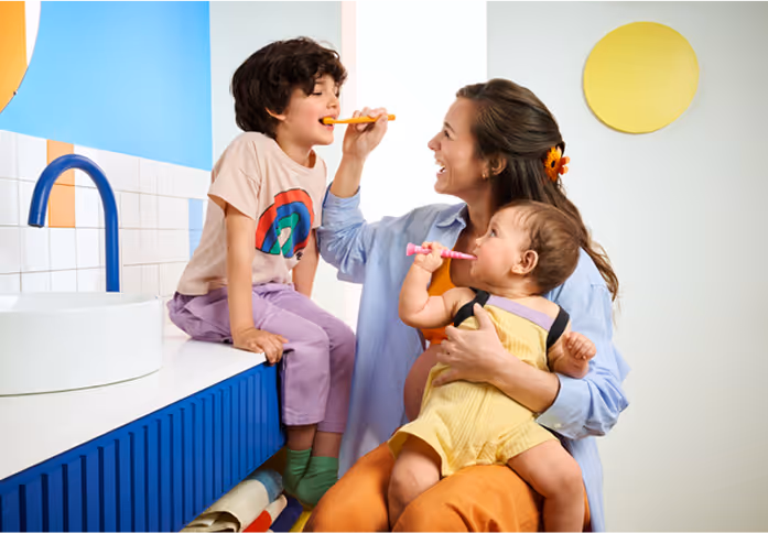 Mother helping two young children brush their teeth in a brightly colored bathroom.