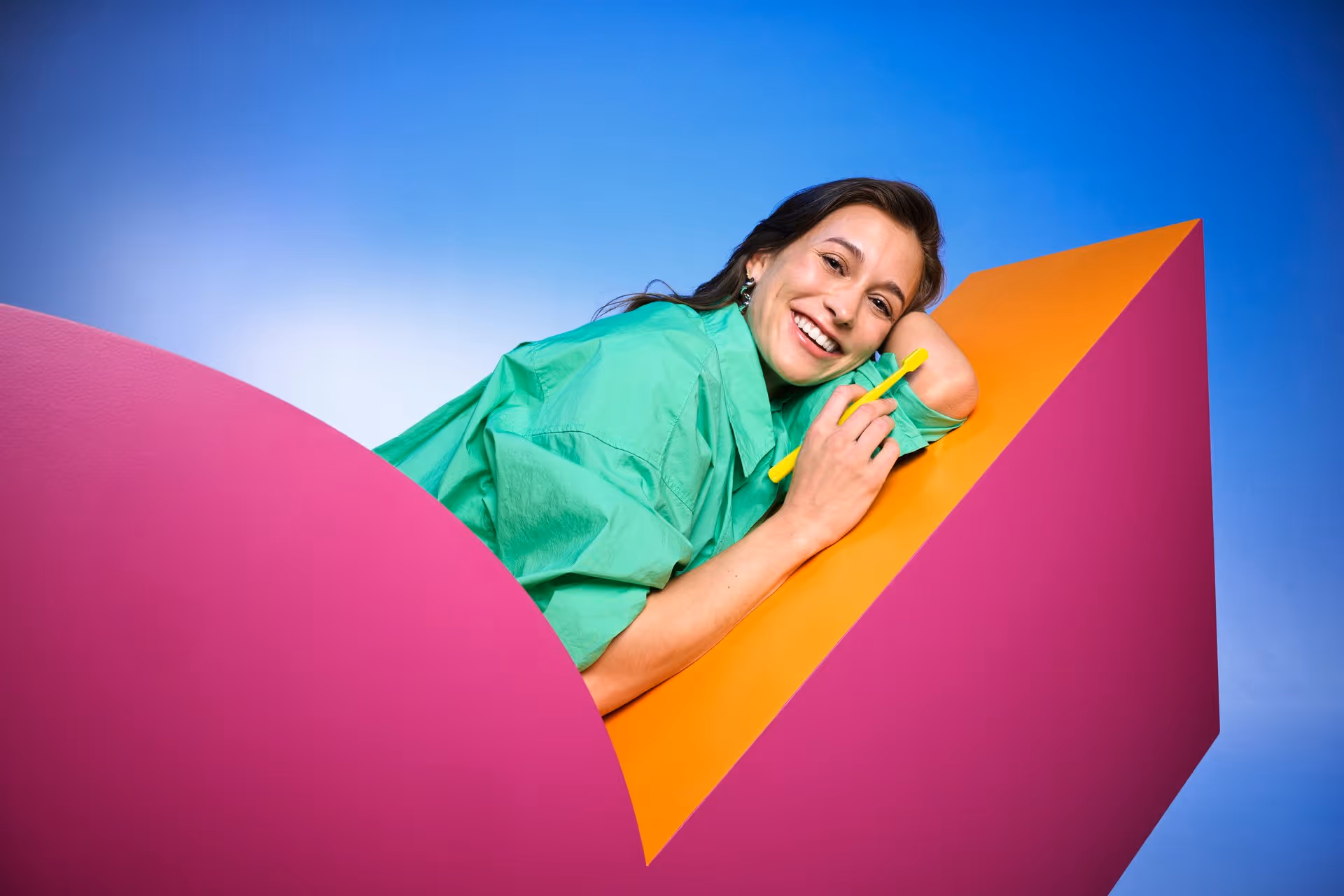 Smiling woman in a green shirt leaning on an orange and pink geometric structure while holding a yellow toothbrush.