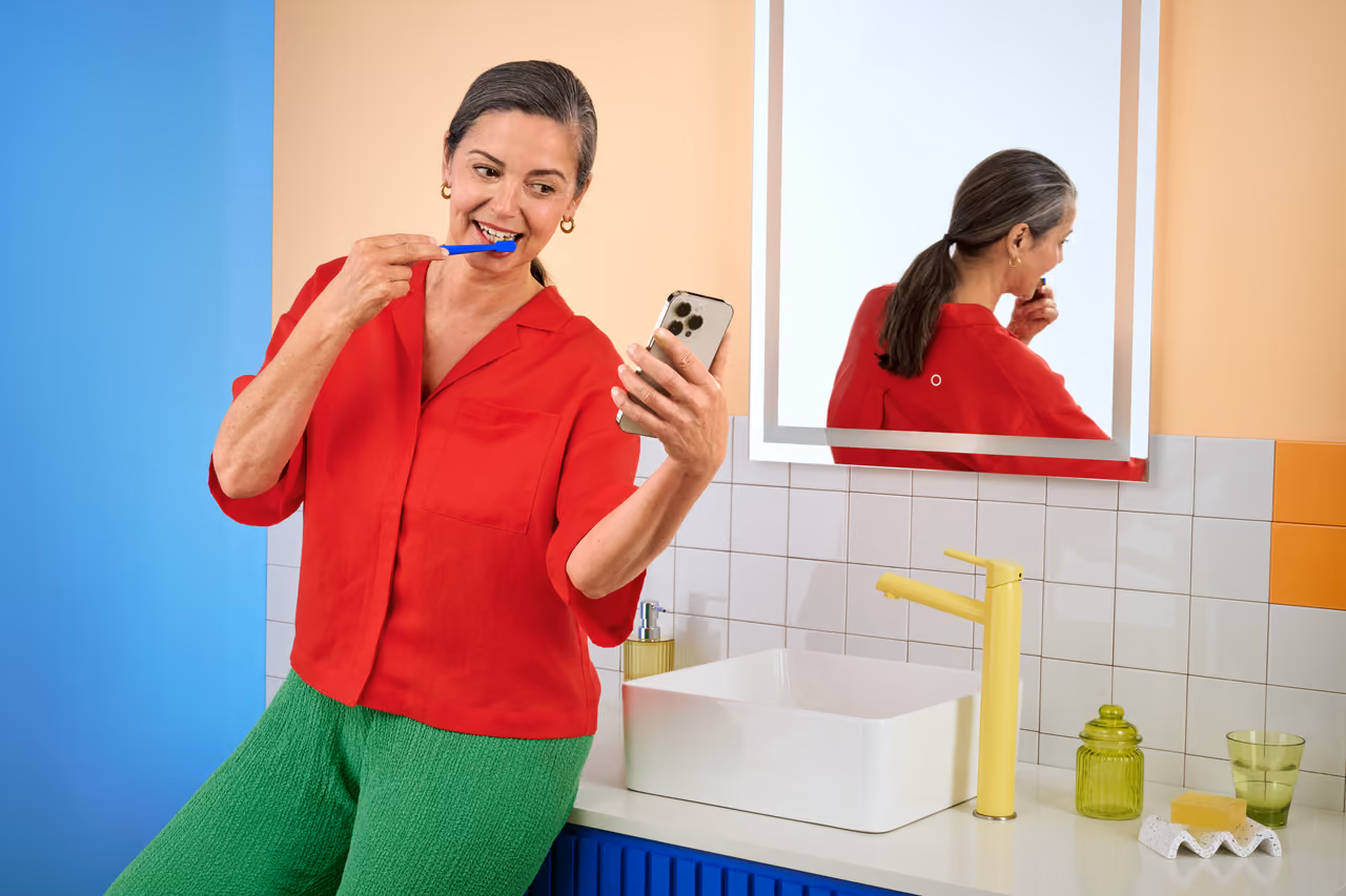 Woman in red shirt brushing her teeth while holding a smartphone in a colorful bathroom with a yellow faucet and white sink.