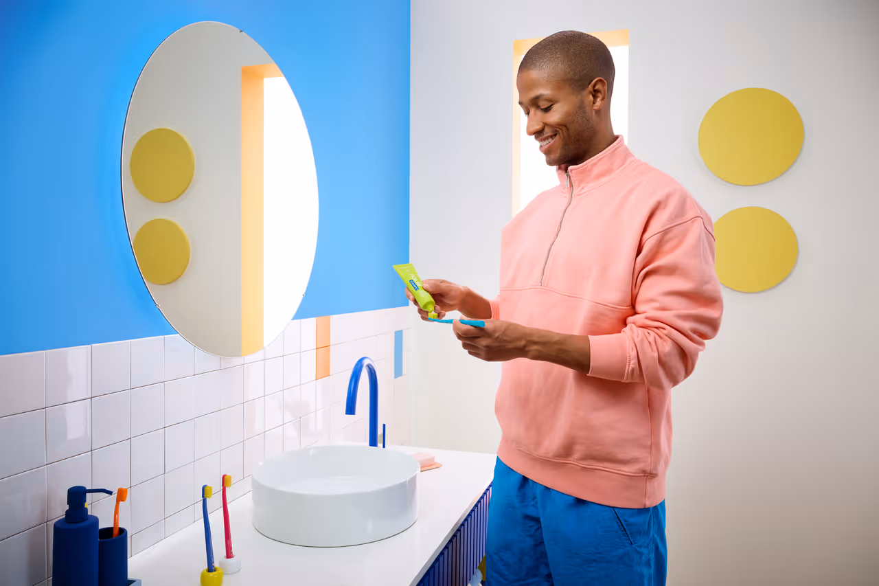 Man in a pink sweatshirt smiling while putting toothpaste on a blue toothbrush in a colorful bathroom.