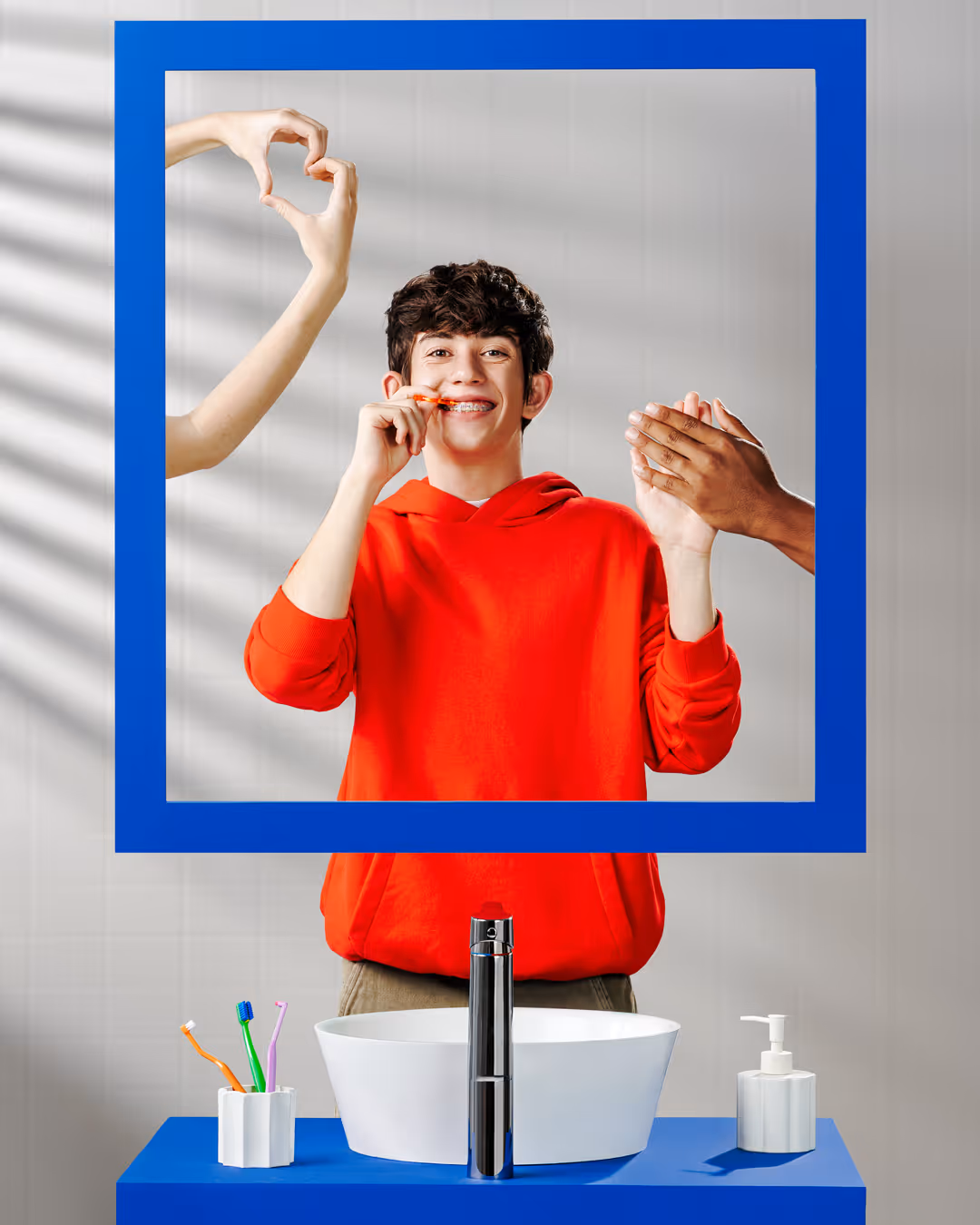 Teen boy with braces brushing his teeth in front of a mirror, framed by a blue square, with hands forming a heart and clapping on either side.
