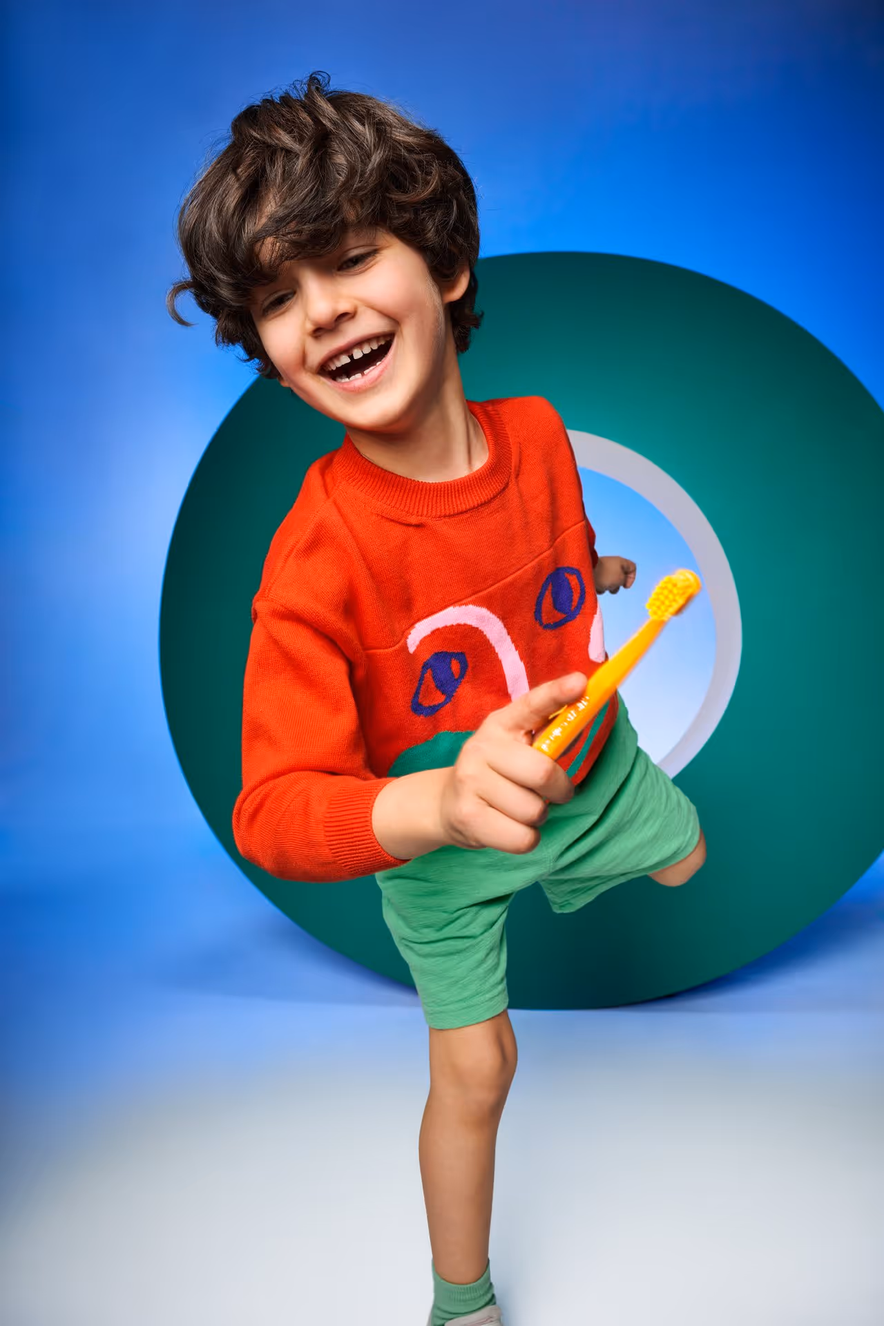Smiling boy in a red sweater and green shorts holding a yellow toothbrush in front of a blue and green circular backdrop.