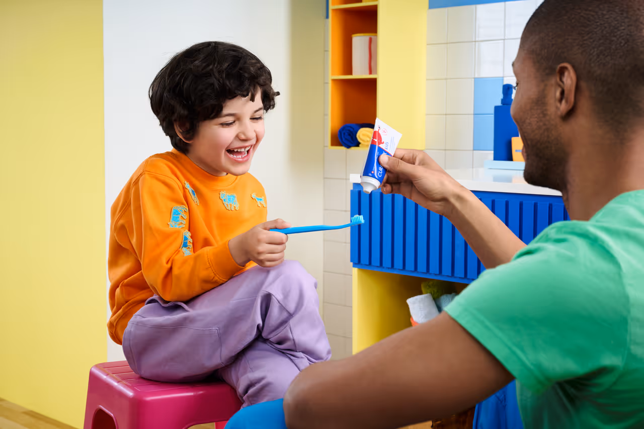 Smiling child sitting on a pink stool holding a blue toothbrush while an adult in a green shirt squeezes toothpaste onto it in a colorful bathroom.