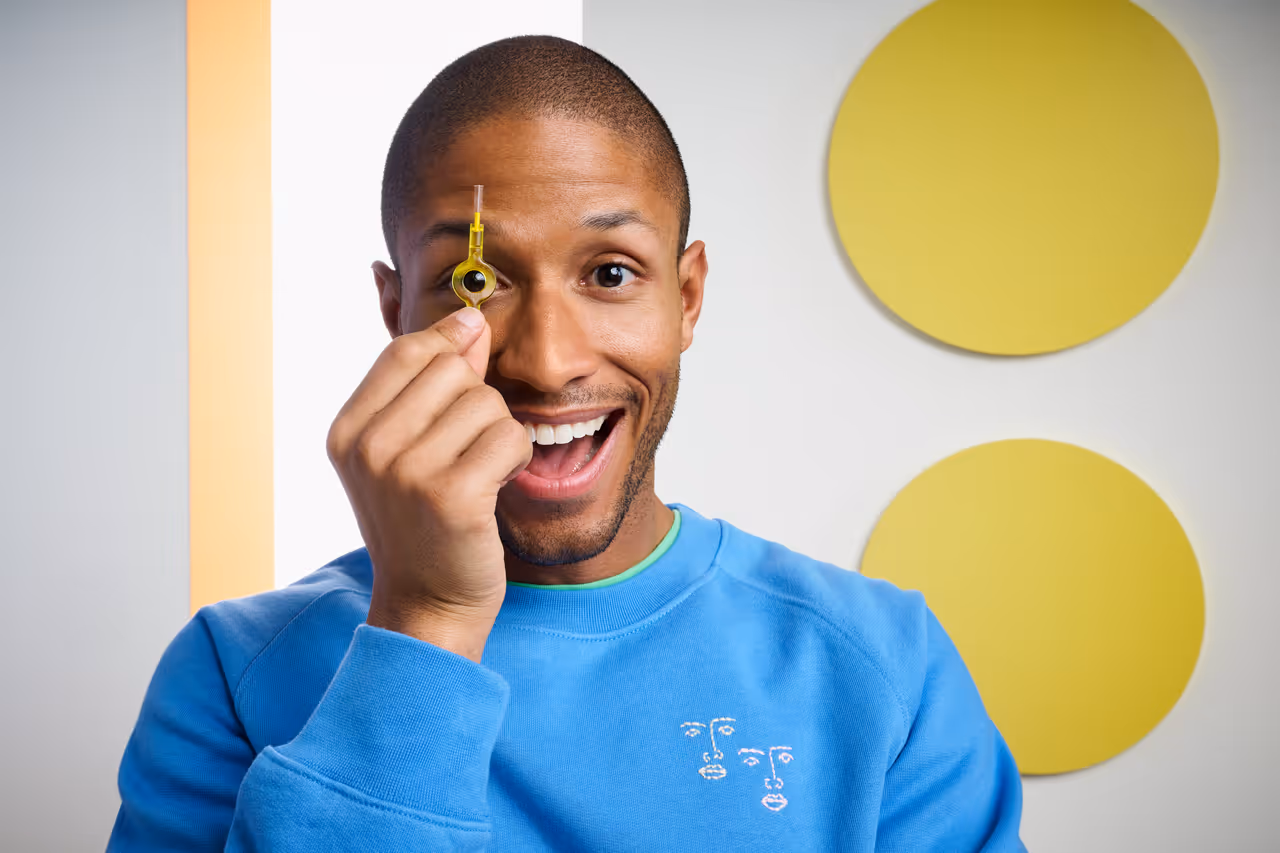 Smiling man in blue shirt holding a yellow interdental brush up to his eye, with two large yellow circles in the background.