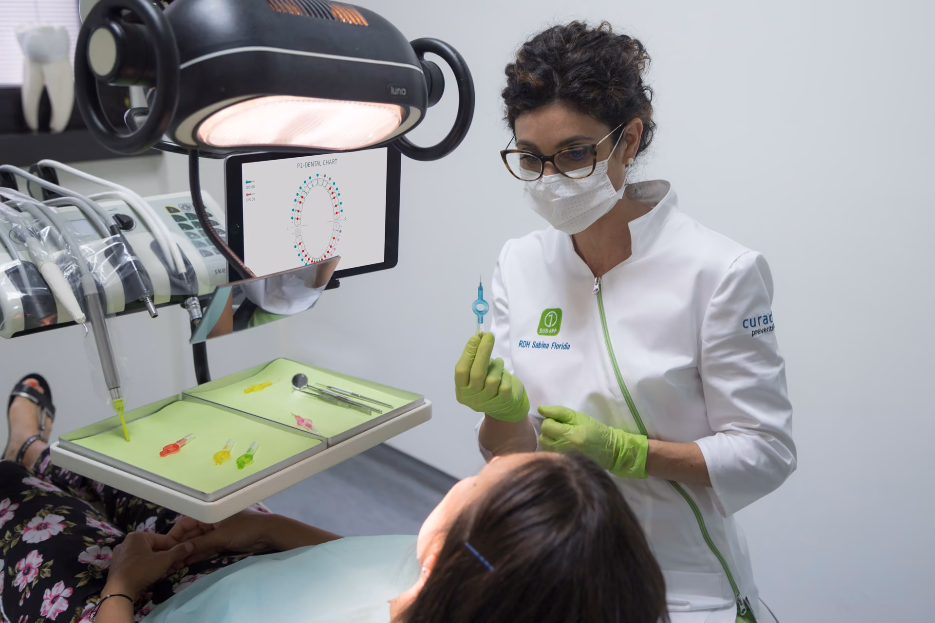 Dentist wearing gloves and mask holding a dental instrument while examining a patient in a clinic.