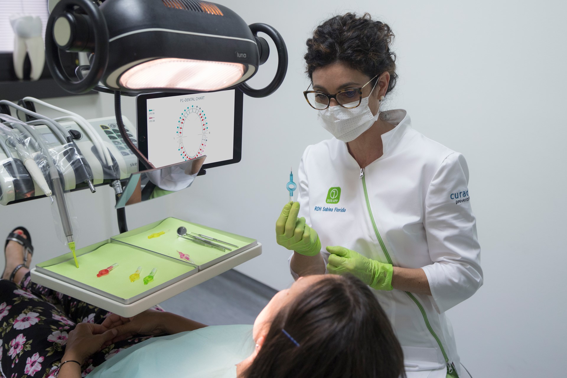 Dentist wearing gloves and mask holding a dental instrument while examining a patient in a clinic.