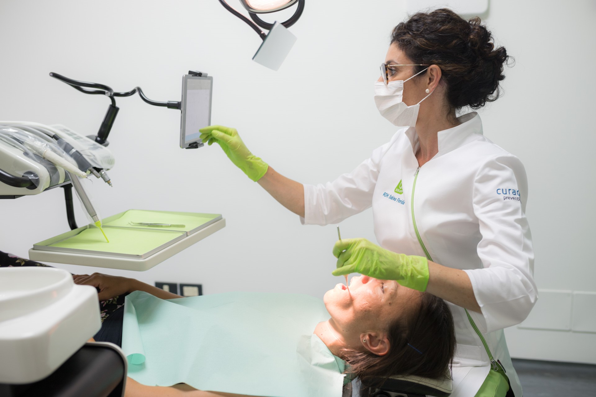 Dentist wearing a mask and green gloves examining a patient's mouth while looking at a tablet in a dental clinic.