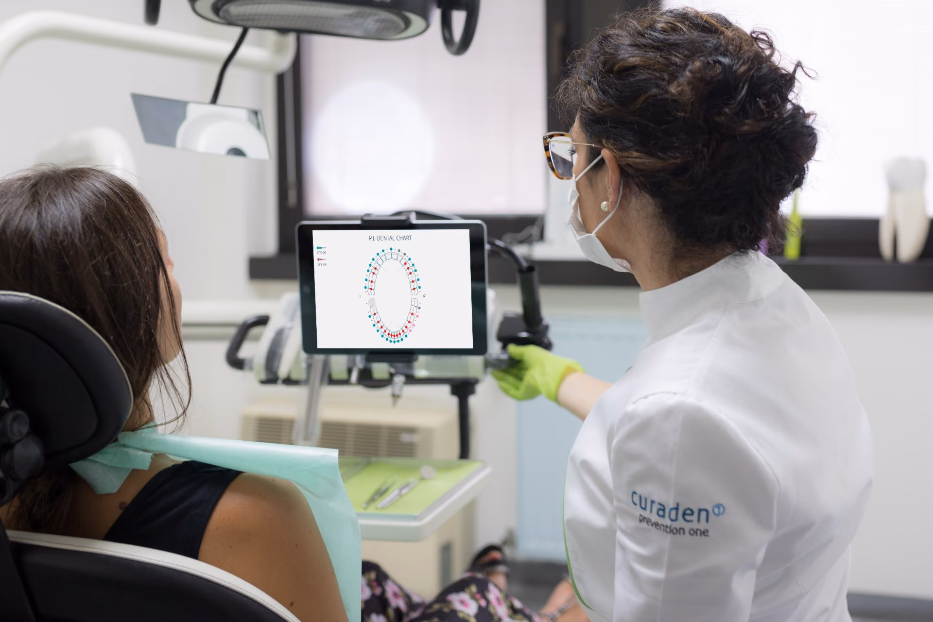 Dentist in gloves and mask showing a dental chart on a screen to a patient seated in a dental chair.