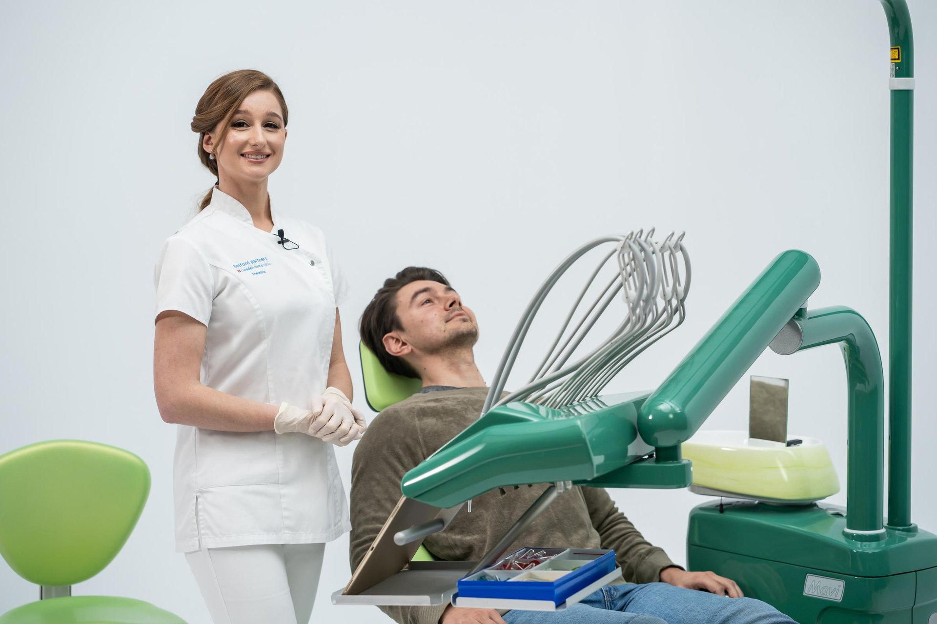Female dentist in white uniform and gloves standing next to a male patient reclining in a dental chair with dental equipment nearby.