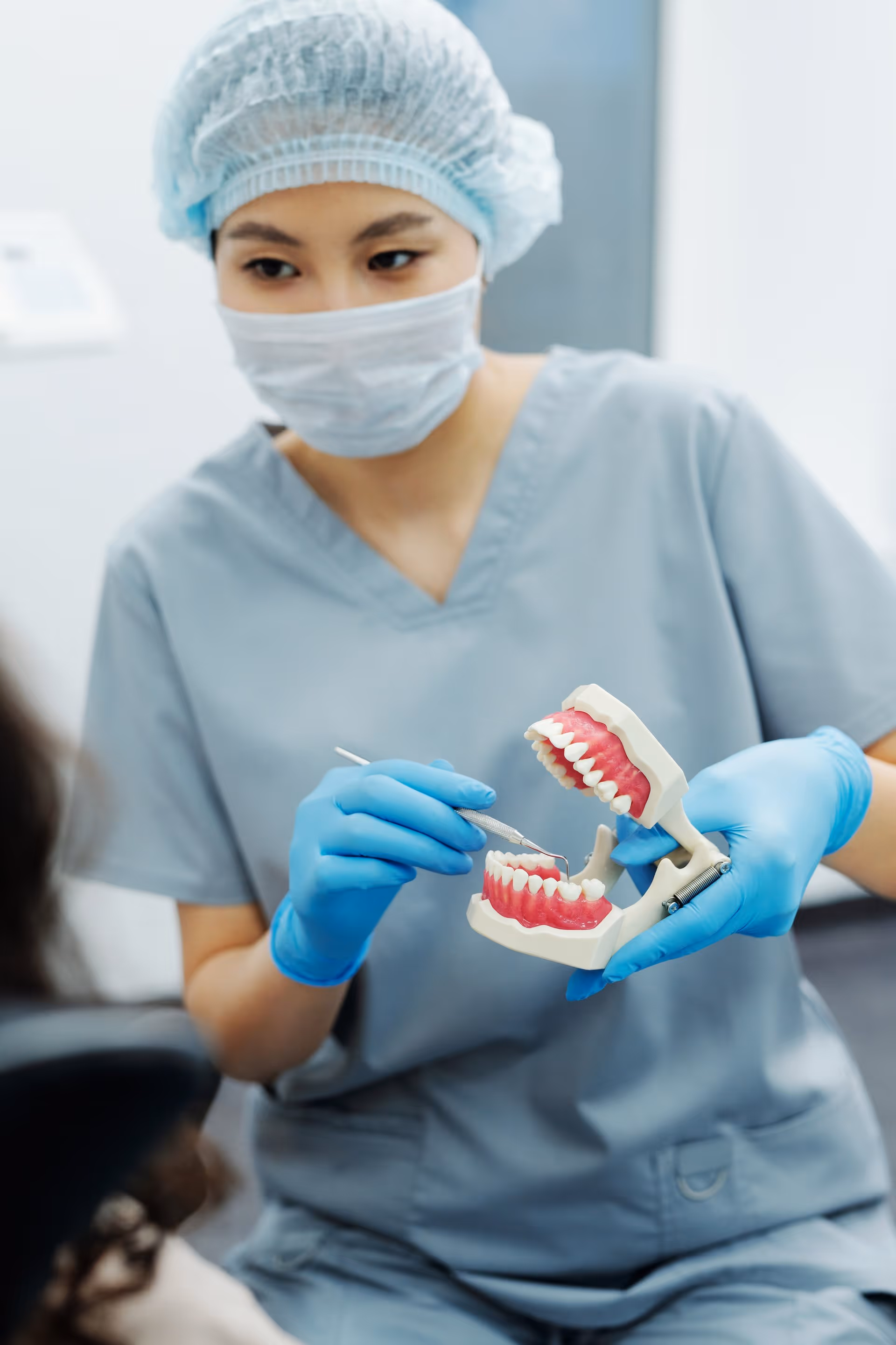 Dentist wearing surgical cap, mask, and gloves demonstrating dental care using a teeth model and dental tool.