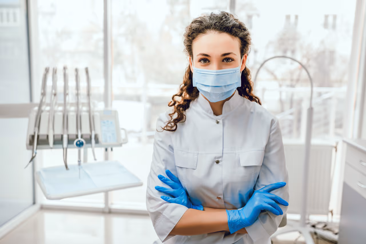 Female dentist wearing a surgical mask and blue gloves with arms crossed in a bright dental clinic.