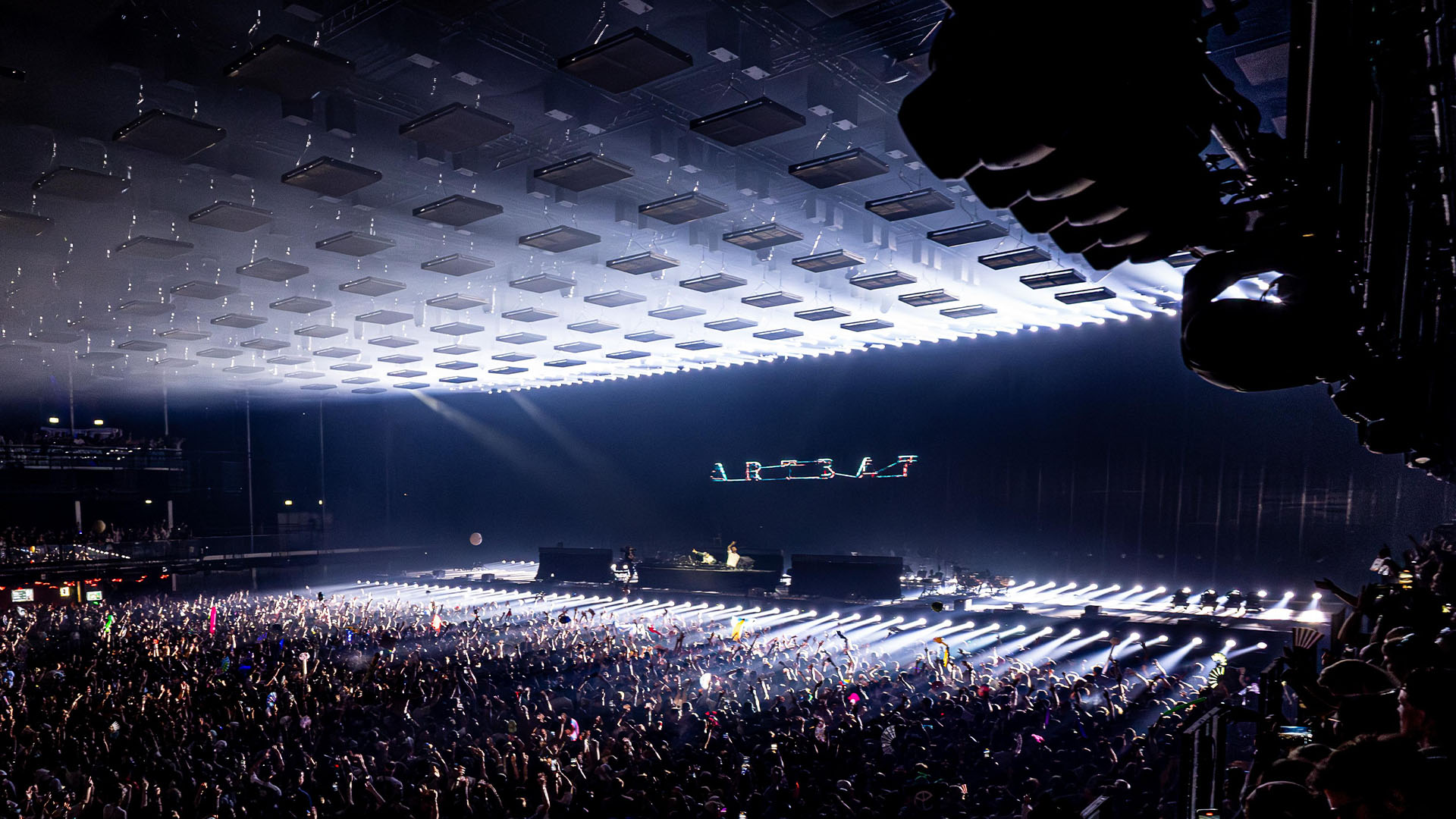 Wide audience view of a concert with a grid of suspended rectangular panels and white stage lighting