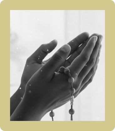 Hands held together in prayer holding a beaded rosary against a light background.