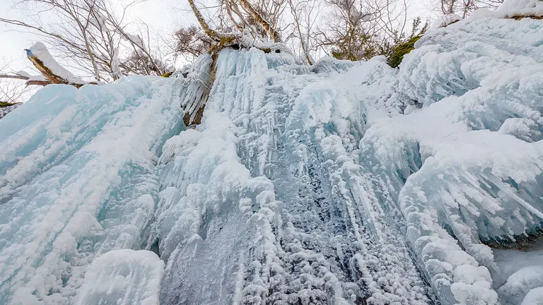 Close-up view of a frozen waterfall with icicles and snow-covered rocks under bare tree branches.