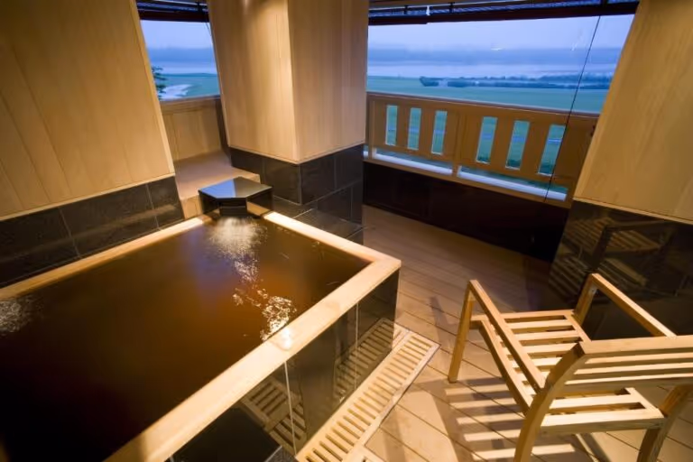 Indoor wooden hot spring bath with water flowing in and a wooden chair on a balcony overlooking a foggy river landscape.