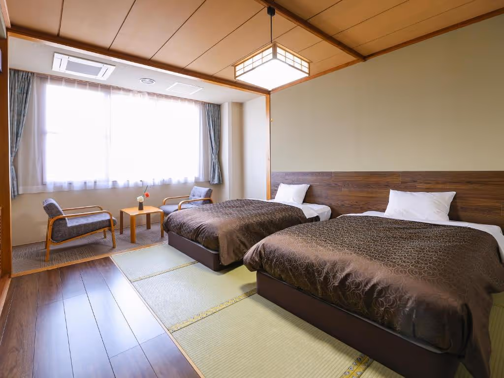 Hotel room with two beds covered in brown patterned blankets, a tatami mat floor, wooden ceiling, and a seating area with two chairs and a small table near a large window.