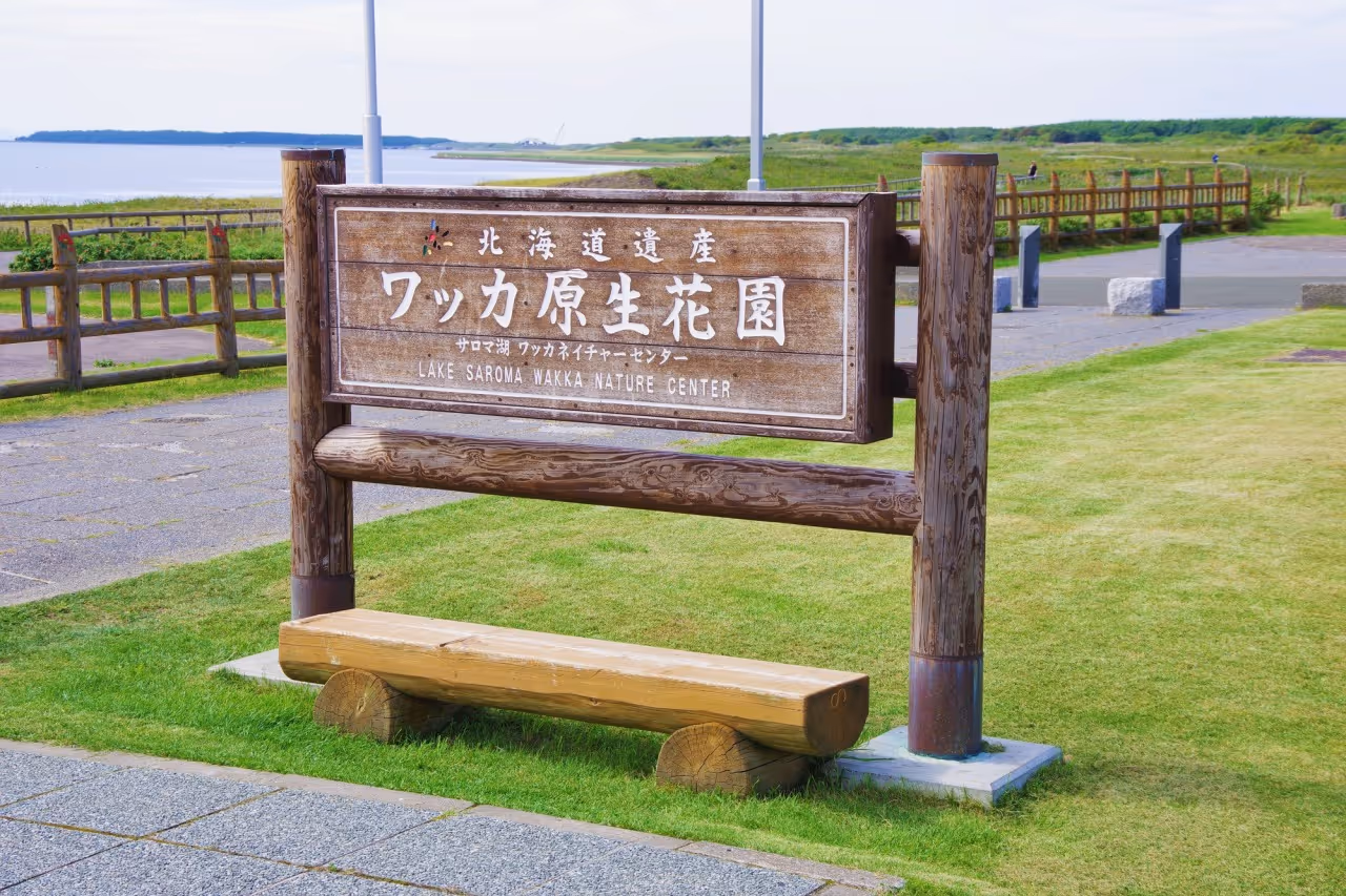 Wooden sign for Lake Saroma Wakka Nature Center on a grassy area near a paved path and a body of water in the background.