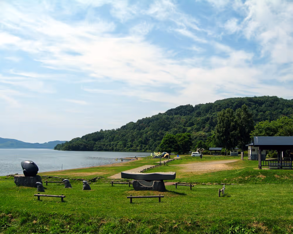 Grassy park area by a lake with sculptures, tents, and forested hills under a partly cloudy sky.