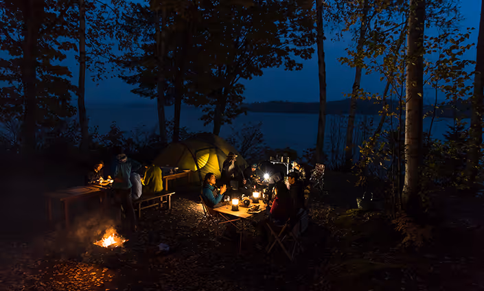 Group of people camping at night around a lit campfire and lanterns with a tent and lake in the background.