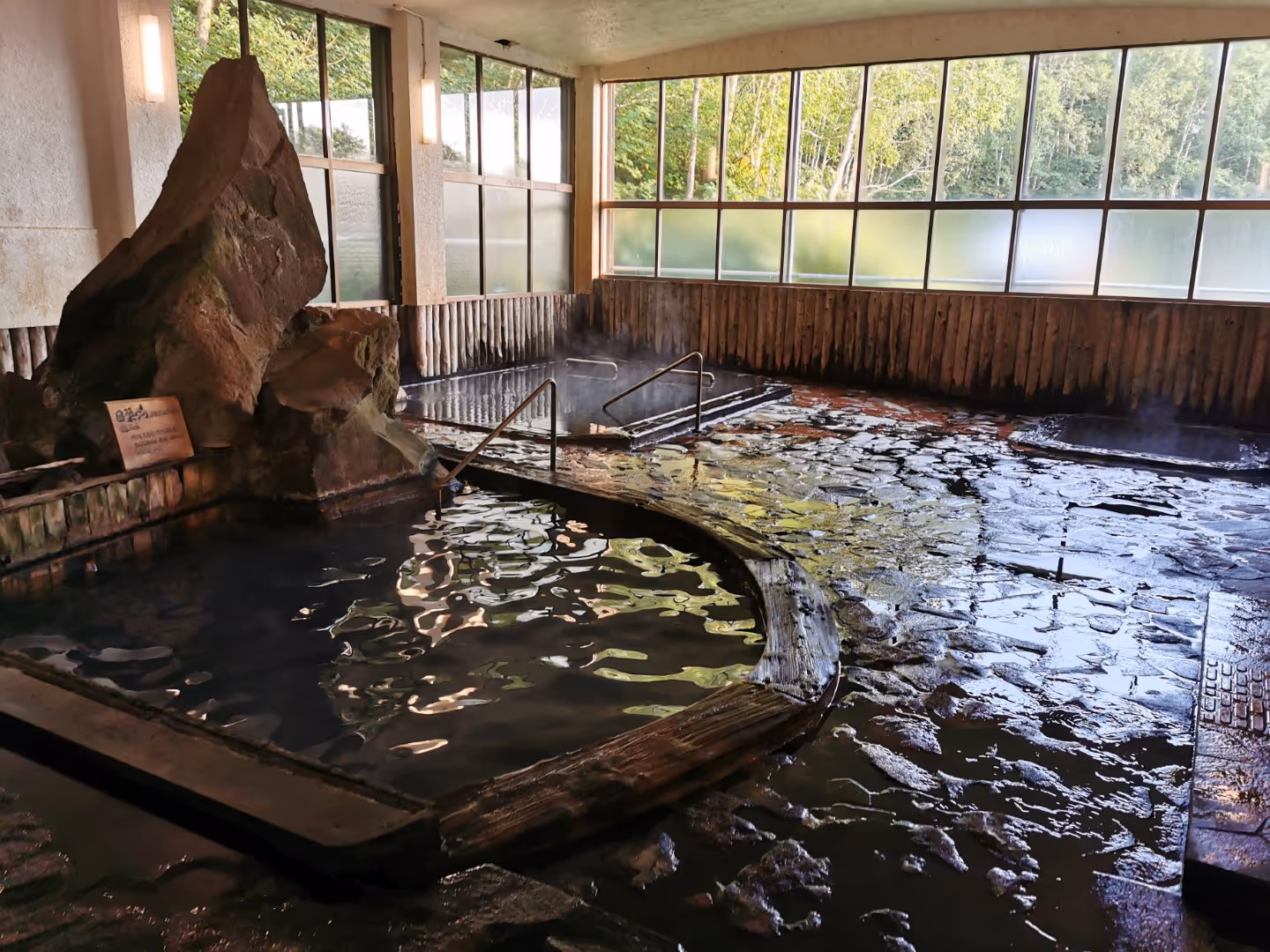 Indoor Japanese onsen with steaming hot spring baths surrounded by stone floors and large windows showing green trees outside.