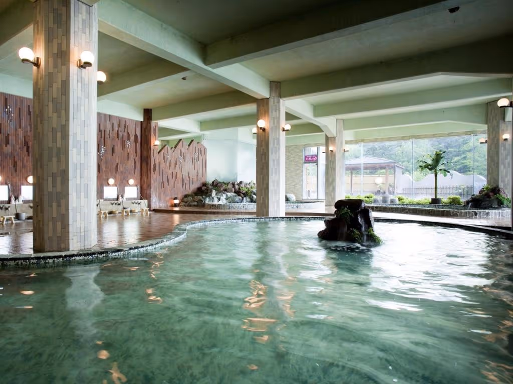Indoor Japanese onsen bath with clear water, tiled pillars, wooden stools along the wall, and large windows showing outdoor greenery.