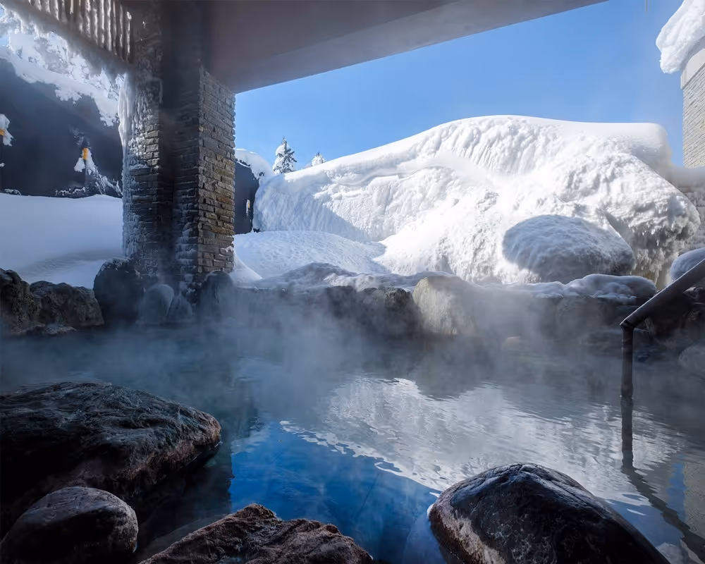 Steaming hot spring bath surrounded by rocks with snow-covered landscape and clear blue sky in the background.