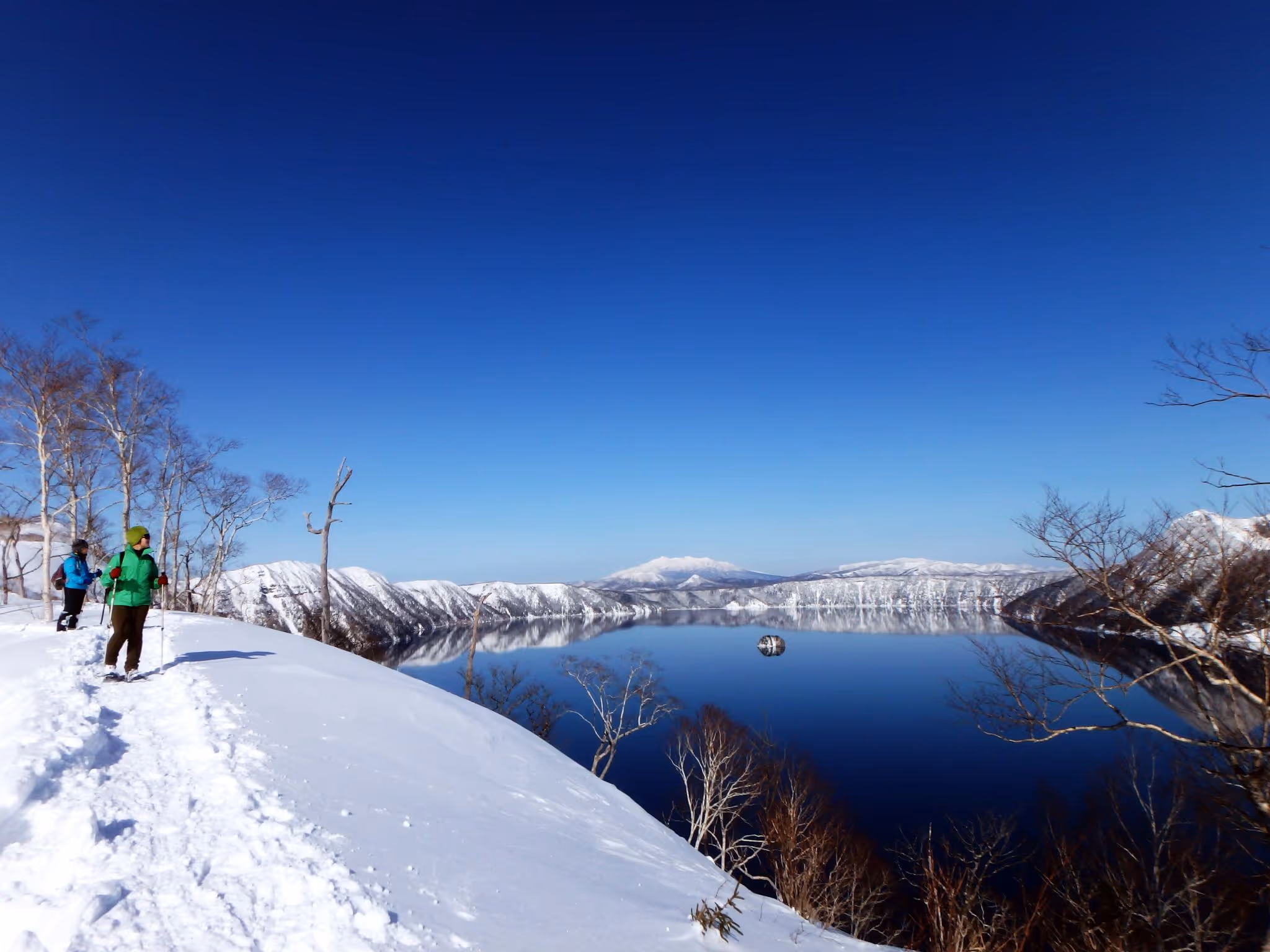 Two hikers stand on a snowy trail overlooking a calm, reflective lake surrounded by snow-covered mountains under a clear blue sky.
