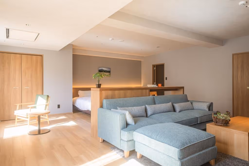 Bright, modern hotel room with a blue sectional sofa, wooden coffee table, armchair, and a partially visible bed behind a wooden divider.