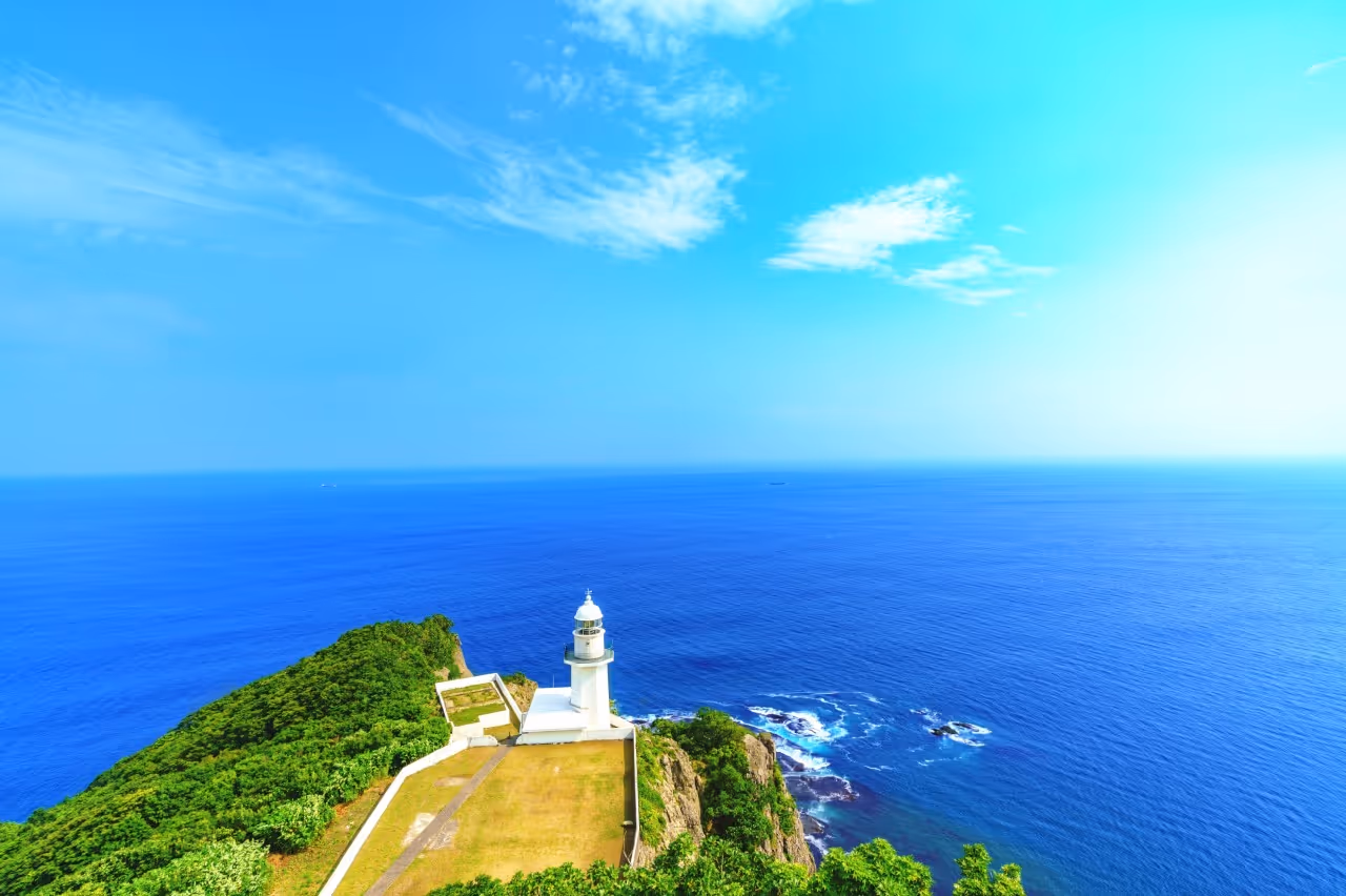 White lighthouse on a green cliff overlooking a vast blue ocean under a clear sky with scattered clouds.