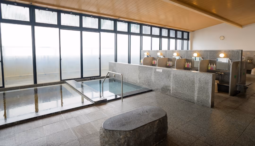 Bright indoor Japanese onsen with two large stone baths, washing stations with mirrors and stools, and large windows along one wall.
