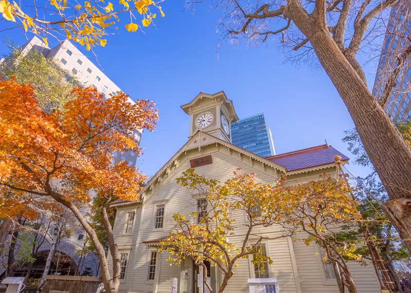 Historic clock tower building surrounded by autumn trees with orange and yellow leaves under a clear blue sky.