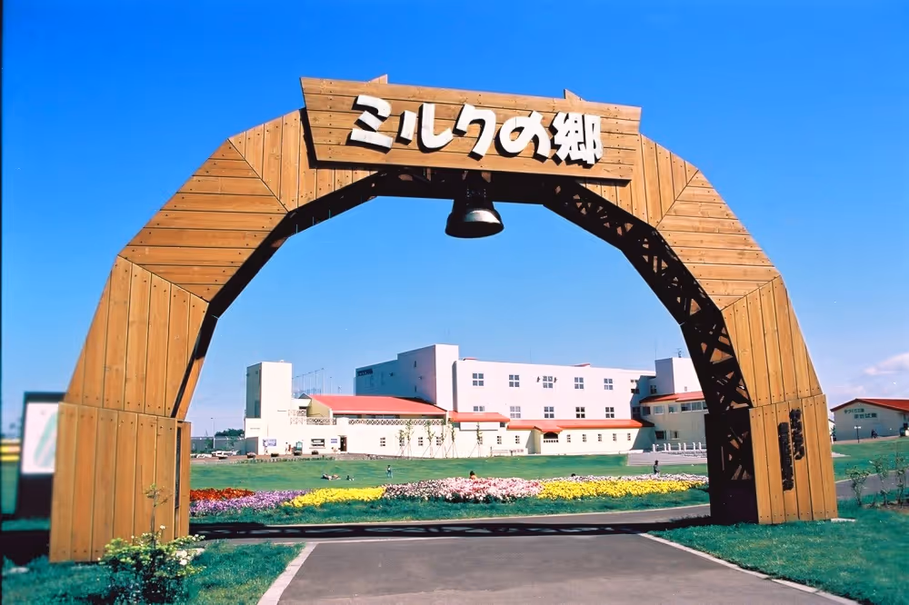 Wooden arch entrance with Japanese text, leading to a large white building with red roofs and colorful flowerbeds under a clear blue sky.