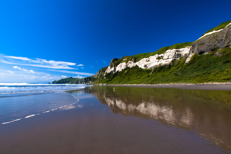 Clear blue sky over a calm beach with waves gently touching the shore and white cliffs reflected in wet sand.