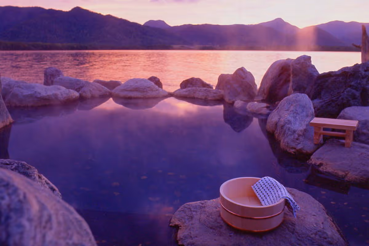 Outdoor hot spring pool surrounded by rocks at sunset with a wooden bucket and towel on a stone.