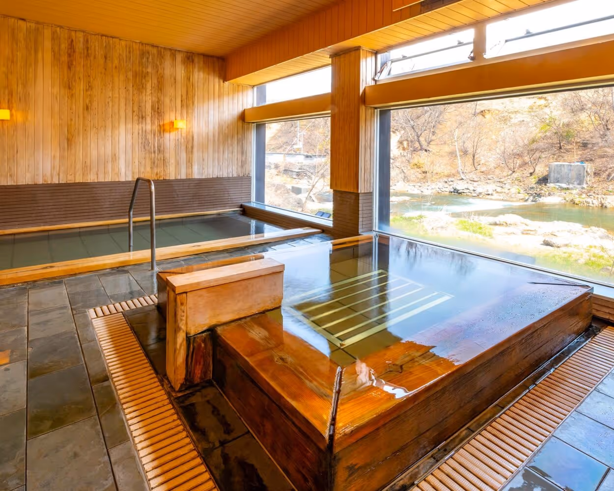 Indoor hot spring baths with wooden frames and large windows overlooking a riverside landscape.