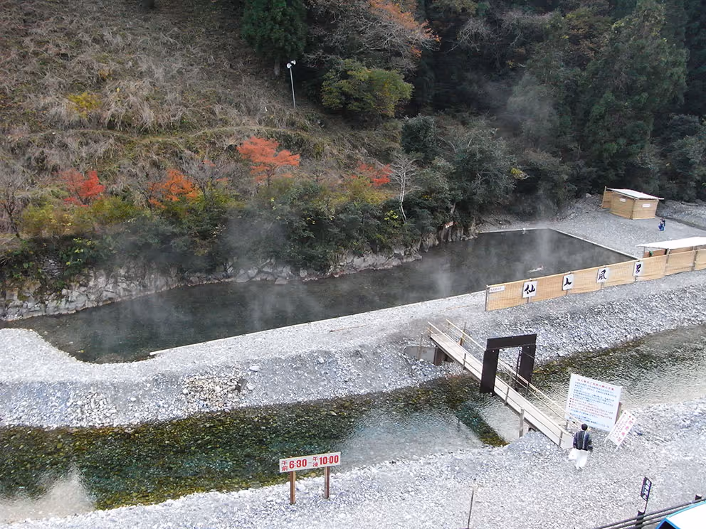 Outdoor hot spring bath surrounded by rocky banks and trees with autumn foliage, with a small wooden bridge crossing a clear river.