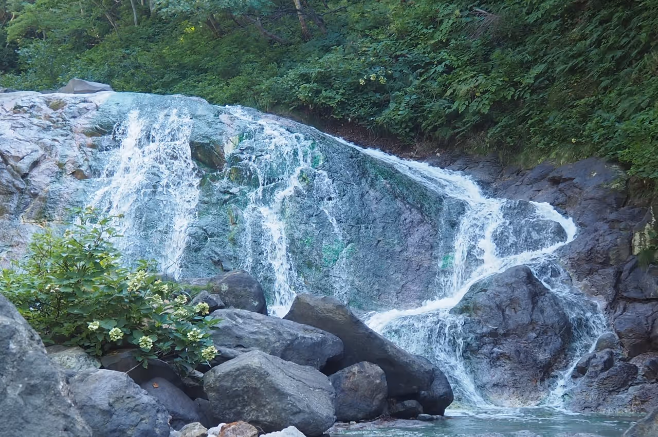 Small waterfall cascading over mossy rocks surrounded by green foliage and large boulders.