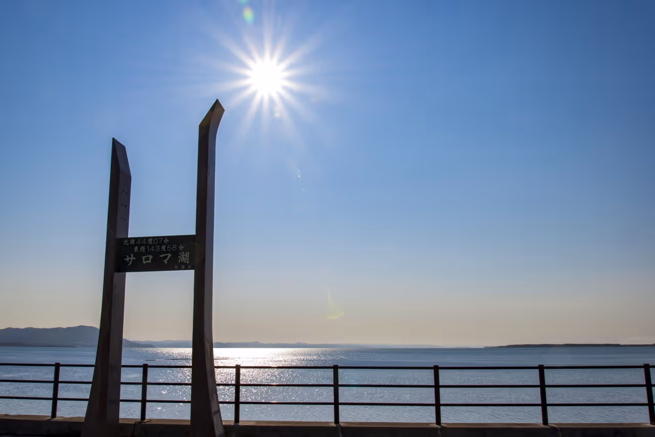 Bright sun shining over calm lake with metal signpost and railing in foreground under clear blue sky.